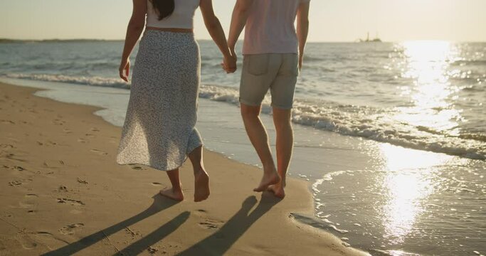 Young loving couple spend time on the beach at sunset. Man and woman enjoying relaxing walk on the beach holding hands. Only legs are taken into the frame. Travel vacation lifestyle.