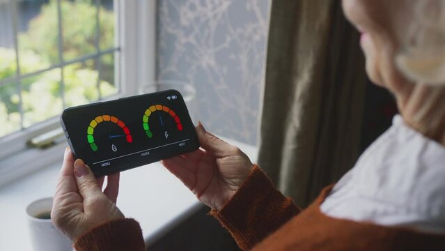 Senior woman standing by window holding smart energy meter looking at consumption of gas and electricity at home during cost of living crisis - shot in slow motion