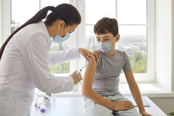 Female doctor makes antiviral vaccine injection for preteen boy in vaccination center. Child in protective mask receives booster dose of covid-19 vaccine. Concept of vaccination of children. - obrazy, fototapety, plakaty