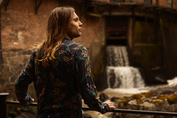 A caucasian man with long hair in a colorful shirt standing infront of a old brick house holding a...