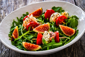 Fresh salad - mozzarella, crudo ham, fig, cherry tomatoes and leafy greens on wooden table
