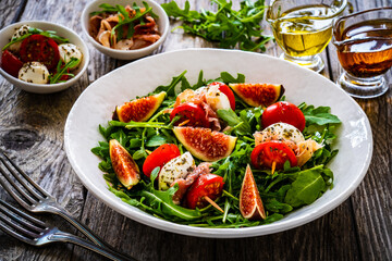 Fresh salad - mozzarella, crudo ham, fig, cherry tomatoes and leafy greens on wooden table
