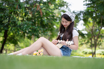 Naklejka na ściany i meble Young Asian woman enjoying and relaxing by playing Ukulele in the park