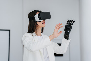Young brunette student woman with disability in vr headset at office raising hands enjoying virtual reality. Girl with bionic hand prosthesis exploring using 3d vision. Electronics, medicine.