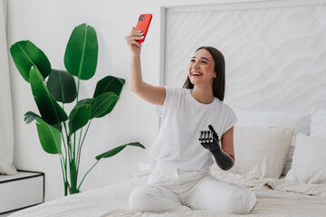 Attractive Spanish girl with bionic arm prosthesis in white clothes  sitting on bed making video...