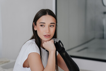 Close up of beautiful Italian brunette girl with disability leaning on bionic mechanical arm prosthesis and another hand. Pretty young woman with cyber arm looking aside sitting on bed, home.