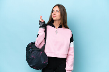 Young sport woman with sport bag isolated on blue background with fingers crossing and wishing the...