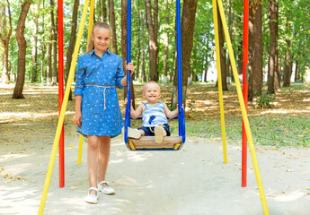 older sister in the park swings her younger brother on a swing on a sunny summer day