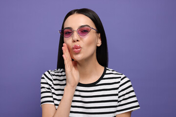 Beautiful young woman in stylish sunglasses blowing kiss on purple background