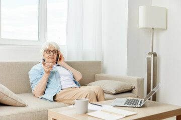 a pleasant elderly woman is sitting at home on the sofa dressed in a blue shirt, with a laptop on the coffee table, having a telephone conversation emotionally gesticulating with her hand