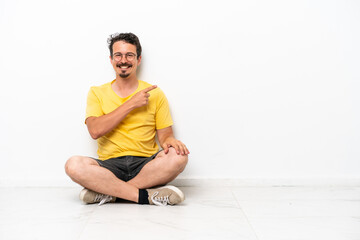 Young caucasian man sitting on the floor isolated on white background pointing to the side to present a product