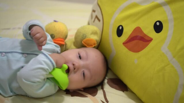 Cute Asian baby chew the teether toy at bed beside yellow duck plush toy