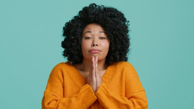 Pleading funny young African American woman 20s years old wears orange sweater hold hands folded in prayer begging about something making wish keep fingers crossed isolated on blue color background 4K