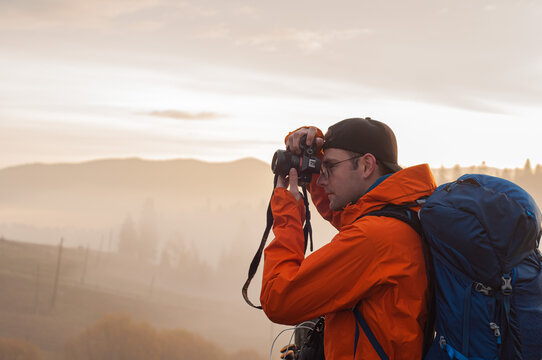 a man with a large tourist backpack takes pictures with a camera at sunrise against the backdrop of mountains and forests landscape tourism travel hiking trekking