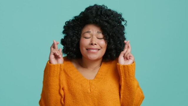 Young pleading cute woman of African American ethnicity wearing orange sweater keeping fingers crossed, praying begging about something making wish isolated on plain pastel blue studio background 4K
