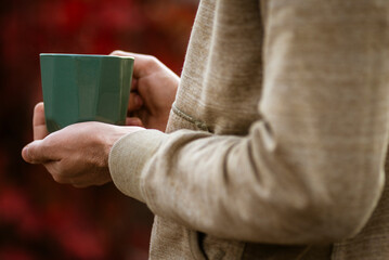 Hands holding green mug  Autumn tea mood red oliage blurred bokeh background outdoors Morning Drinking  Fall cozy concept, backlit vacation  blanket.