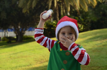 Portrait of a cute boy in a Santa Claus hat, palm trees in the background. The child holds a white jar of drinking yogurt in his hands. The boy drinks yogurt, kefir. Healthy drink during the holidays.