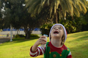 Portrait of a cute boy in a Santa Claus hat, palm trees in the background. The child holds a white jar of drinking yogurt in his hands. The boy drinks yogurt, kefir. Healthy drink during the holidays.