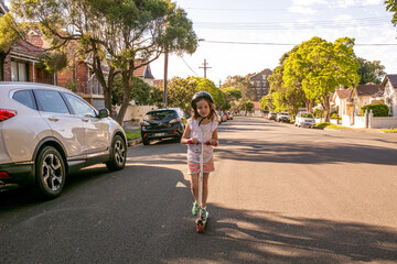 girl riding on a scooter with a helmet on the road with parked cars on a sunny day