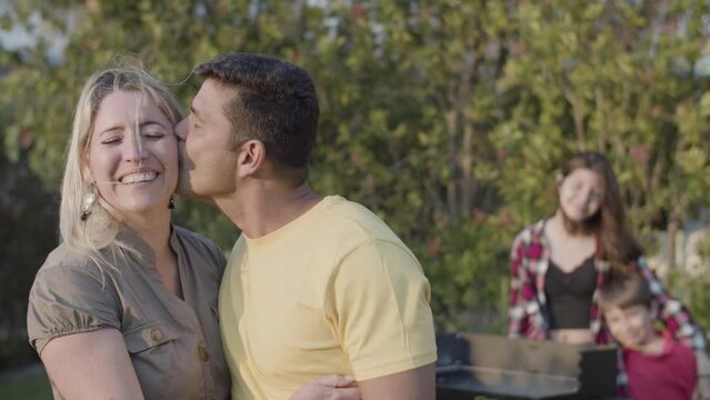 Portrait of happy husband kissing his wife outdoors, their children standing at barbecue grill in background. Family at barbecue party and relationships concept