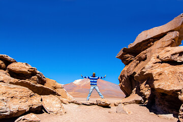 Man with outstretched arms in the desert, surrounded by red rocks with a volcano in the background