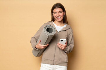 Woman in sport hoodie holding rolled mat and phone on brown background after online workout