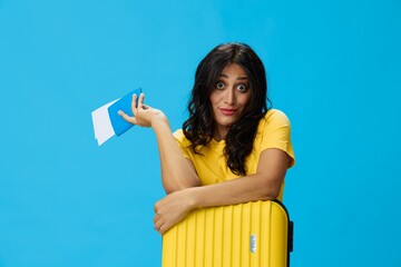 Travel woman with yellow suitcase, passport and ticket in hand, paper plane, in yellow T-shirt on blue background, happiness from travel, glasses, copy space