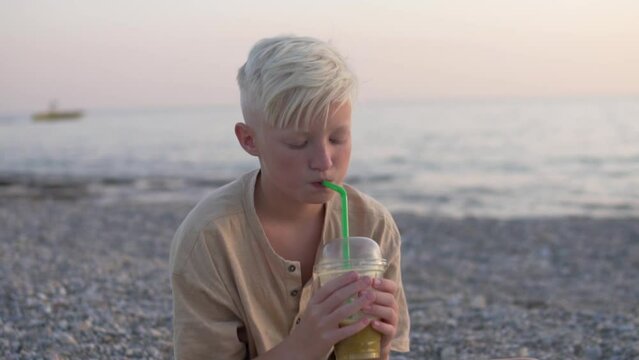 A handsome blond boy drinks freshly squeezed juice on the beach by the sea.