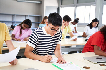 Student taking notes while studying in high school. Satisfied young man while sitting at desk in classroom. Portrait of college guy writing while completing assignment. High quality photo - obrazy, fototapety, plakaty
