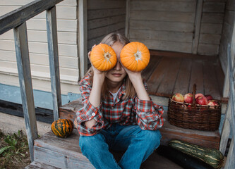Girl is sitting on steps of an old village house with pumpkin in hands near baskets with zucchini and apples. Harvest. Autumn. Fall. Halloween. Thanksgiving day