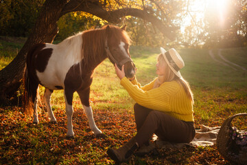 woman and spotted brown pony at sunset in forest, beautiful girl in autumn clothes loves her horse, concept of kindness, animal care, nature and friendship. hat