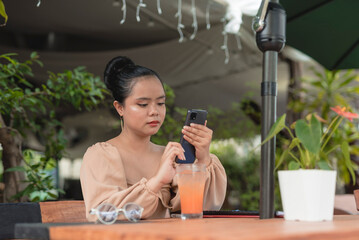 A serious looking Asian woman sits in an al fresco restaurant and browses through the internet with her cellphone on a hot afternoon.