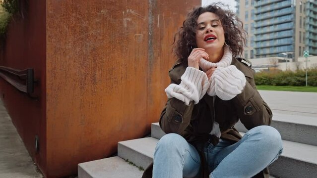 Charming young woman with curly hair joyful relaxed girl in autumn outfit trendy coat pullover smiles into camera sitting on stairs near decorative metal rusty wall street, urban style fashion female