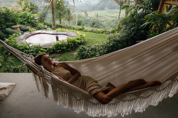 Young beautiful woman lies in a hammock in a bamboo eco-house in the jungle against the backdrop of mountains and a rice field. Ecotourism in Bali