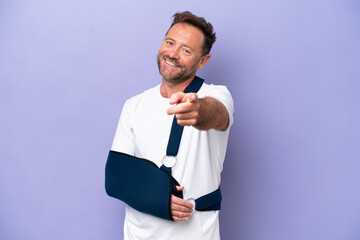 Middle age caucasian man with broken arm and wearing a sling isolated on purple background pointing...