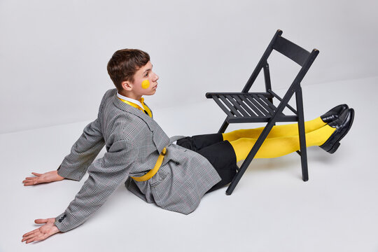 Portrait of stylish teen boy posing in jacket, blazer and yellow tights with weird makeup isolated over grey background