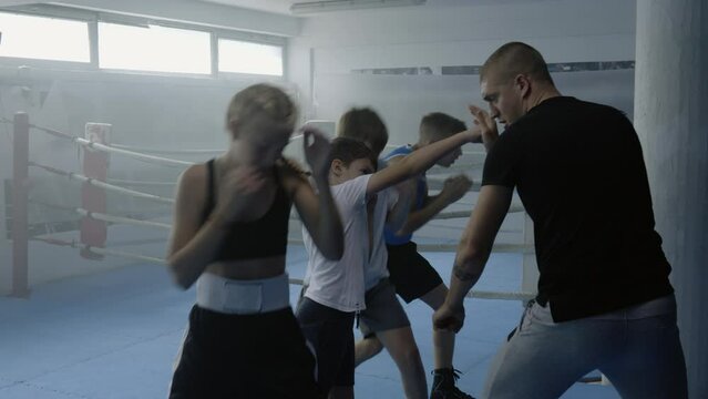 Boxing teacher showing kids punches technique at fighting class, teenagers group practicing kicks in gym. Boys and girls workout, male boxer training children in combat sport club