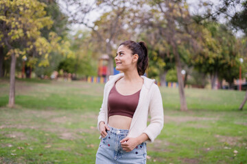 Smiling girl in the park wearing a short maroon blouse, a white jacket and a denim skirt, looking to the side.