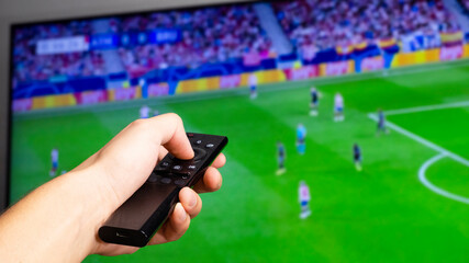 Man hand holding a black tv controller watching a football match on a tv screen in the background