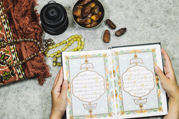 lantern and open Holy Quran with Arabic calligraphy meaning of Al Quran and prayer mat , rosary muslim and bowl dates fruits and woman hands holding a holy book 