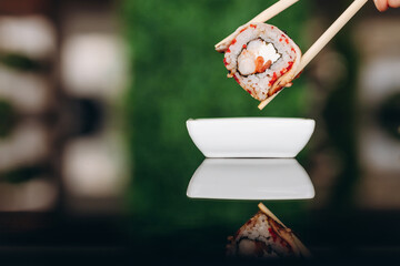 Man's hand picking up sushi with chopsticks and dipping in soy sauce on green background.