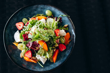 Summer salad bowl with grilled sweet peach, soft cheese, walnuts and fresh arugula on black kitchen table, top view.