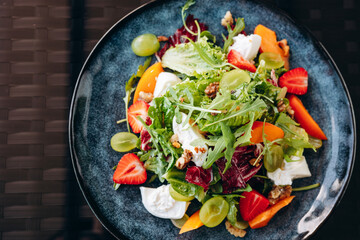 Summer salad bowl with grilled sweet peach, soft cheese, walnuts and fresh arugula on black kitchen table, top view.