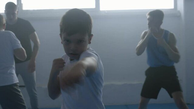 Boy training boxing strike, looking at camera, handheld close up shot. Kid exercising in gym portrait, child practicing at fighting class