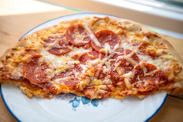 Macro closeup of fresh thin crust half pizza on white plate with melted mozzarella cheese, pepperoni meat slices of unhealthy greasy food