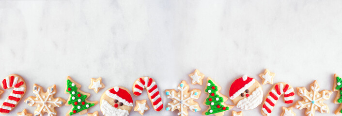 Cute Christmas cookie bottom border. Top view on a white marble banner background with copy space. Holiday baking concept. Santa Claus, tree, snowflakes, candy canes, stars.