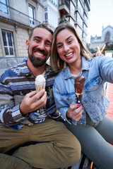 Photo of happy caucasian couple eating ice cream at the city, taking a selfie looking at camera.