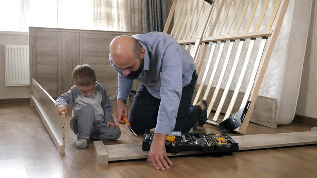 Dad and son are on the floor assembling parts for a new bed in the living bedroom. Father screwing the screws with a screwdriver on the background of a set of tools. Family activity together.