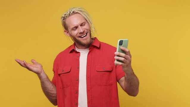 Young blond man with dreadlocks wear red shirt white t-shirt get video call using mobile cell phone do selfie talk conducting pleasant conversation greet with hand isolated on plain yellow background