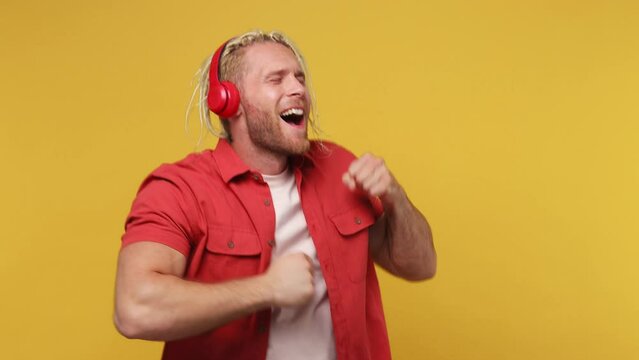 Young vivid cheerful blond man with dreadlocks 30s wear red shirt white t-shirt listen music in headphones dance fooling around have fun gesticulating hands relax isolated on plain yellow background